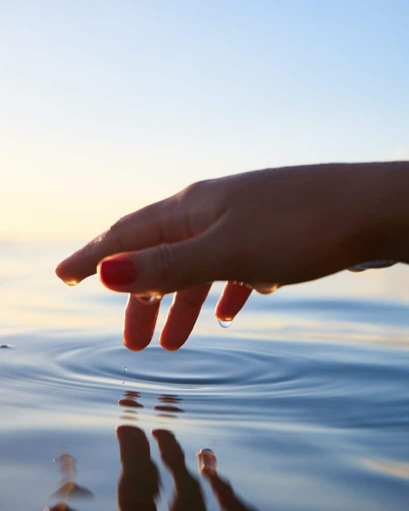 Hand outstretched over calm water with water droplets on the bottom of the hand with sunshine highlighting the image.